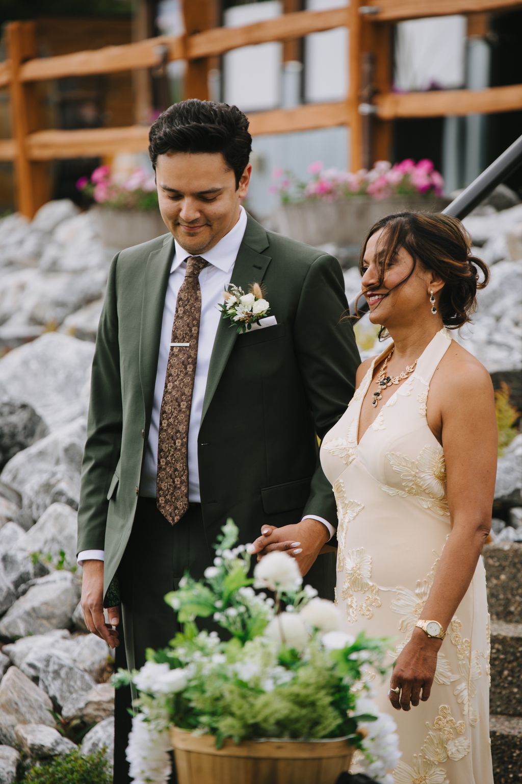 Groom in olive suit and patterned tie with boutonniere smiles beside bride in ivory lace dress, holding hands among white floral arrangements and wooden gard…