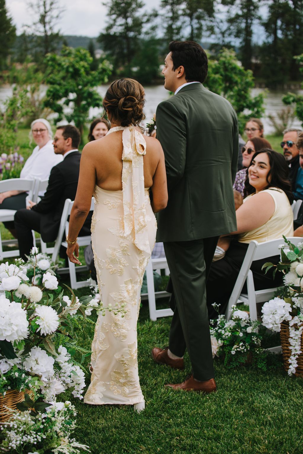 Bride in cream embroidered halter gown and groom in sage suit stand together on manicured lawn overlooking water