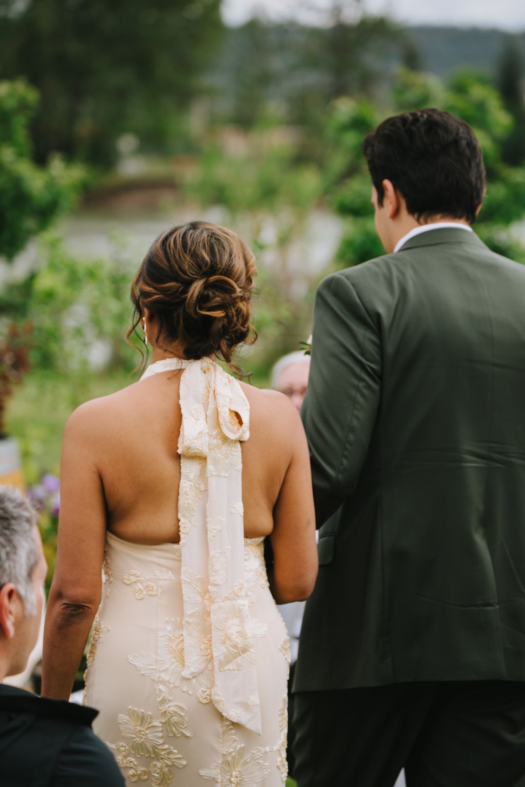 Bride in embellished halter gown with upswept hair stands beside groom in olive suit, gazing toward blurred garden backdrop
