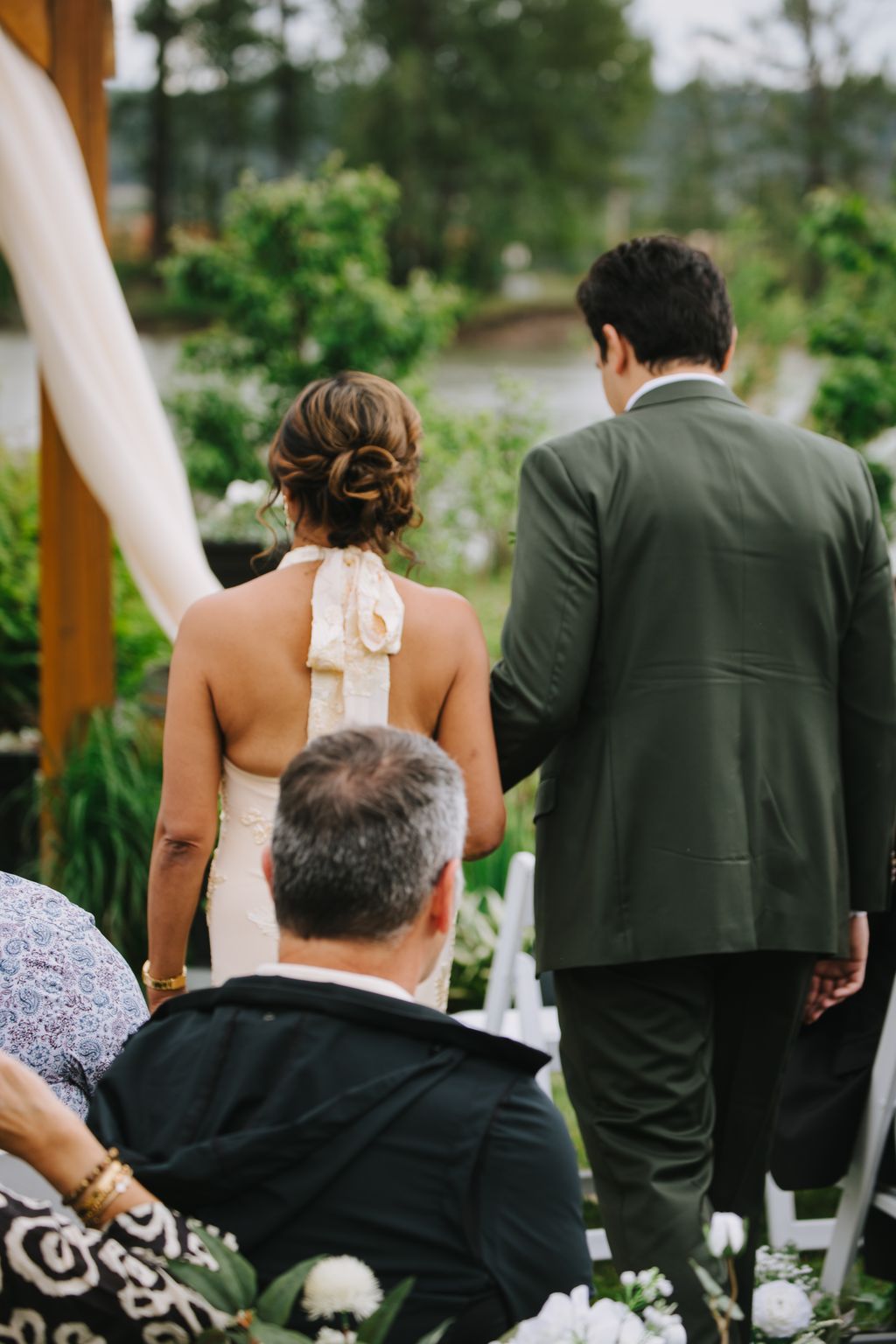 Bride and groom walk together through a lush garden ceremony space with guests in foreground, soft natural light filtering through green foliage