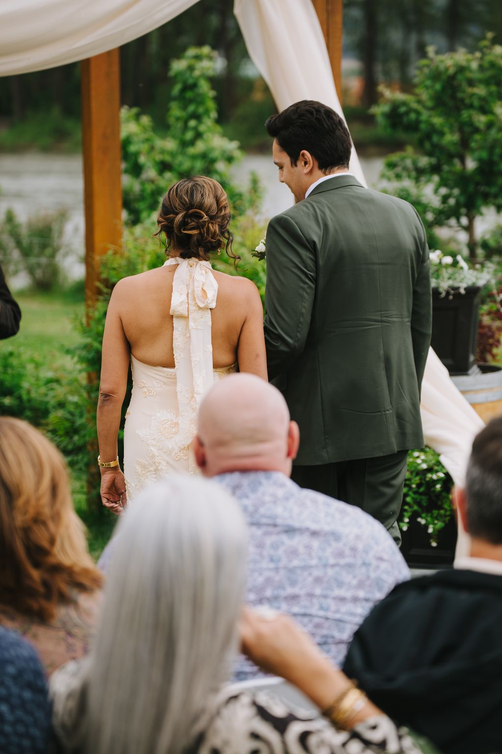 Bride and groom stand together under a draped arbor in an outdoor garden setting, with a guest visible in foreground