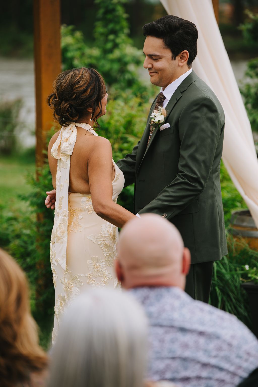 Bride in cream embroidered gown and groom in olive suit exchange vows outdoors with lush greenery behind them