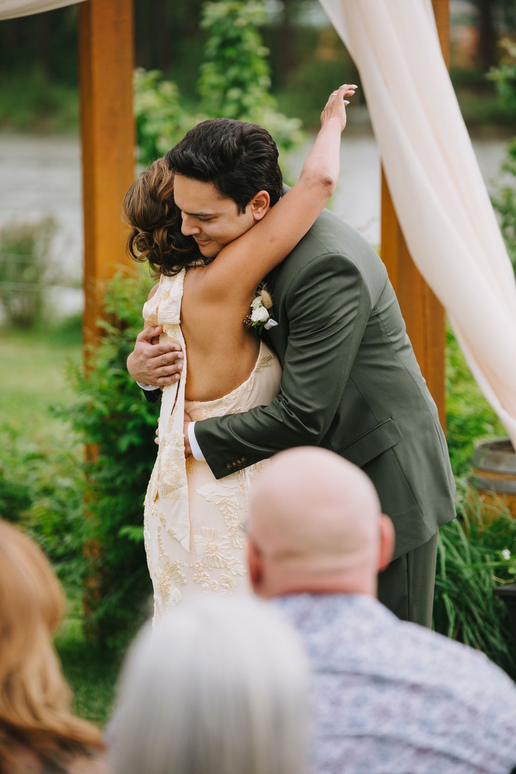 Groom embraces bride from behind in outdoor garden setting, bride''s arm raised joyfully, wearing embellished white dress, soft natural light filtering throug…