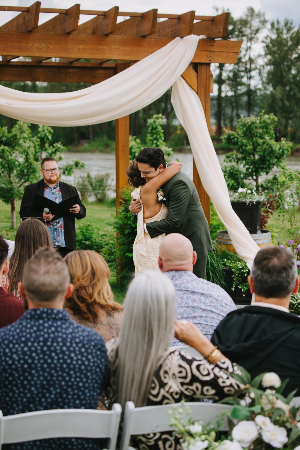 Groom embraces bride under flowing white draping on wooden pergola at outdoor garden ceremony