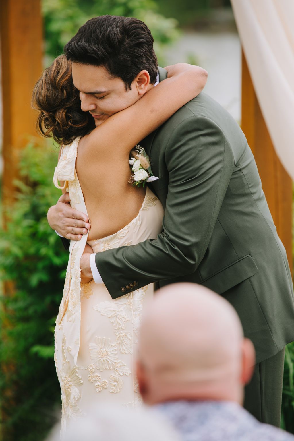 Groom in olive suit embraces bride in embroidered cream gown with corsage, both smiling with eyes closed in tender moment outdoors