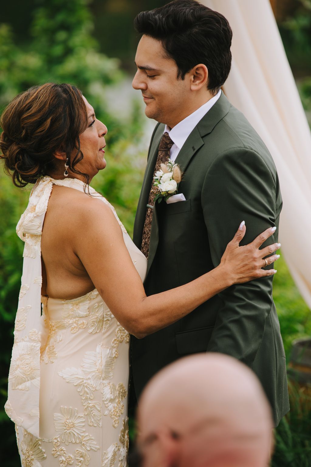 Bride and groom face each other outdoors, smiling warmly in soft natural light with lush green foliage behind them