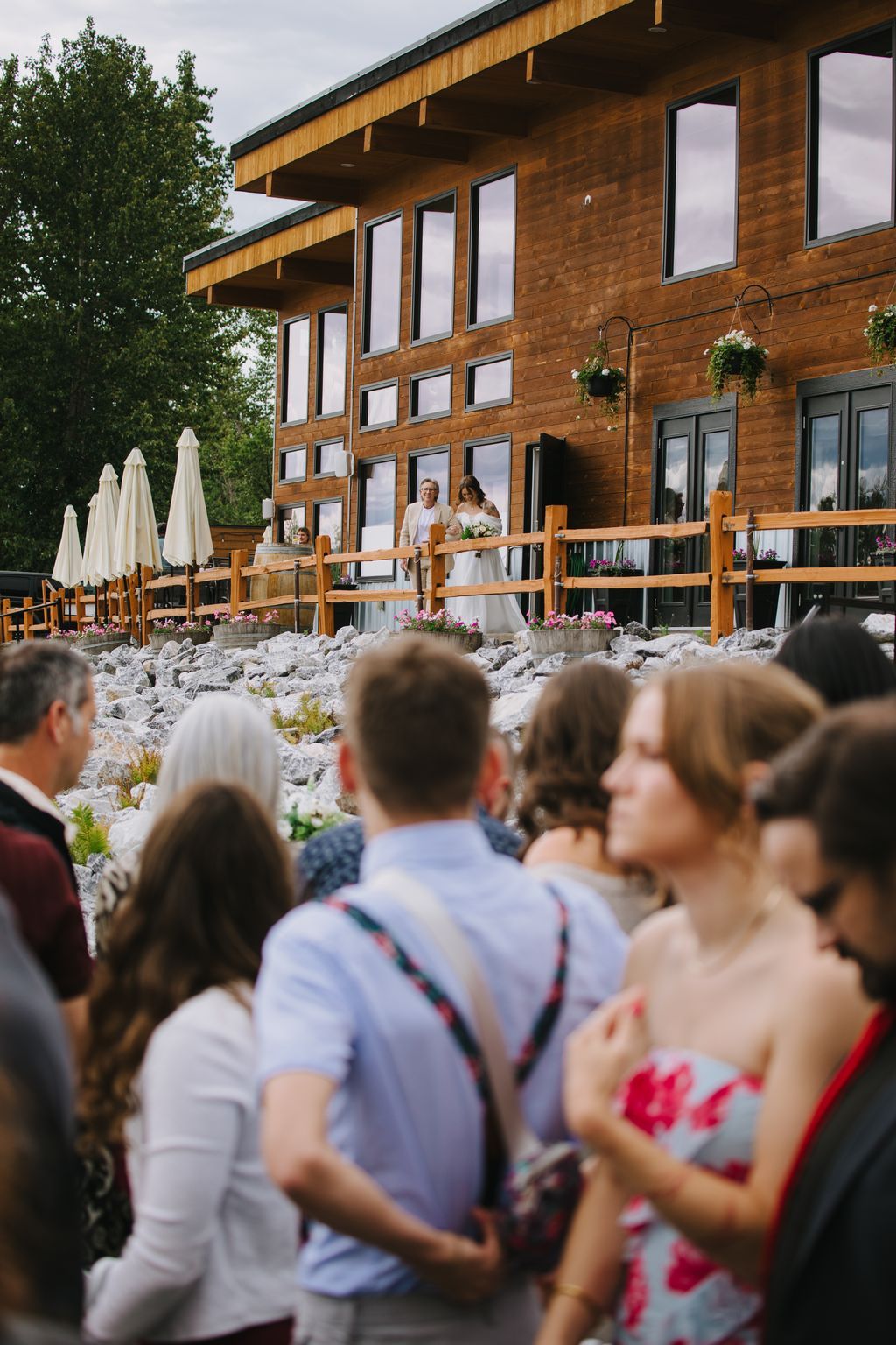 Bride and groom stand on a modern wooden deck while guests gather below on white gravel in bright daylight