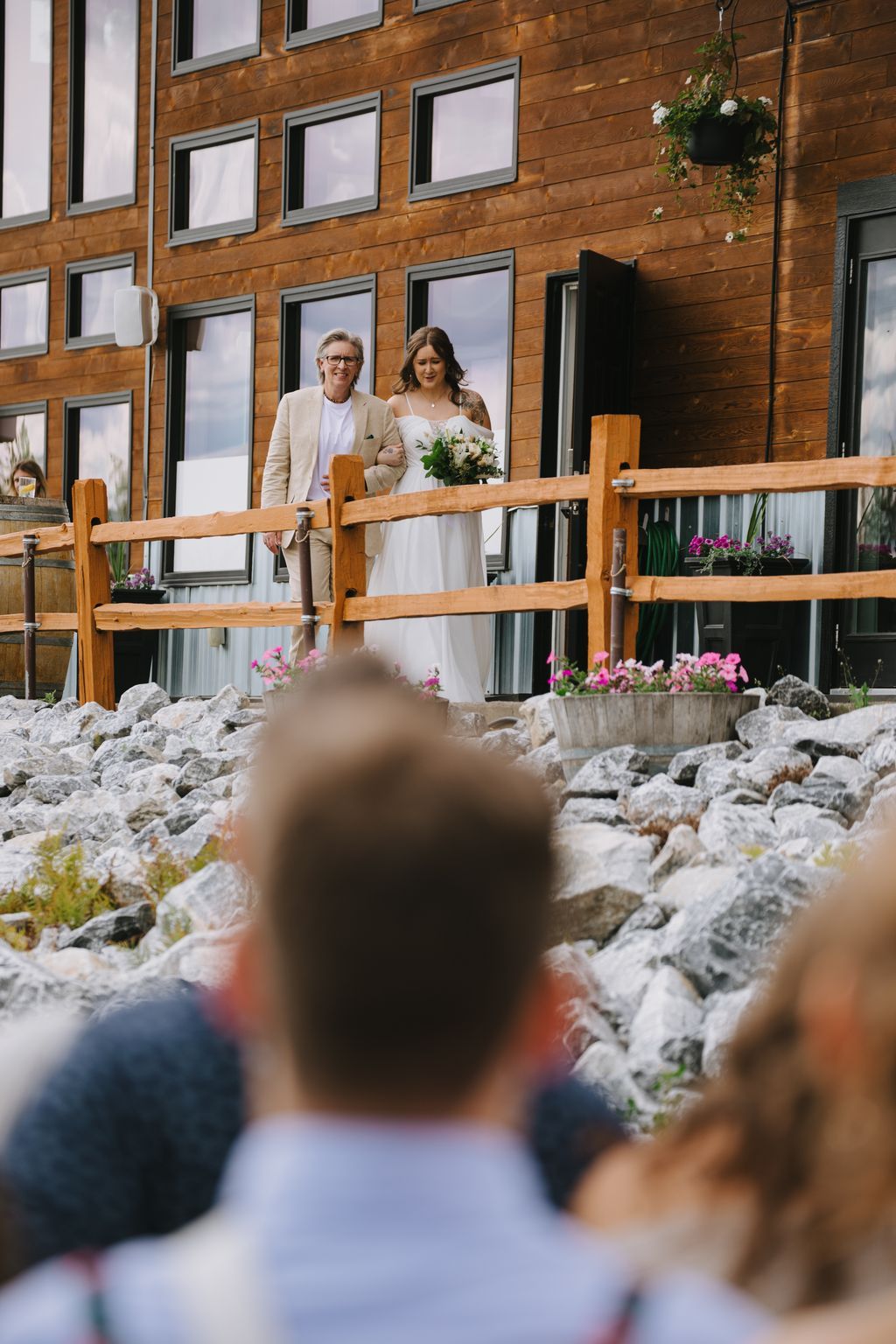 Bride and groom stand on a wooden deck railing against a modern wood-clad building, bride holding white bouquet, soft natural light