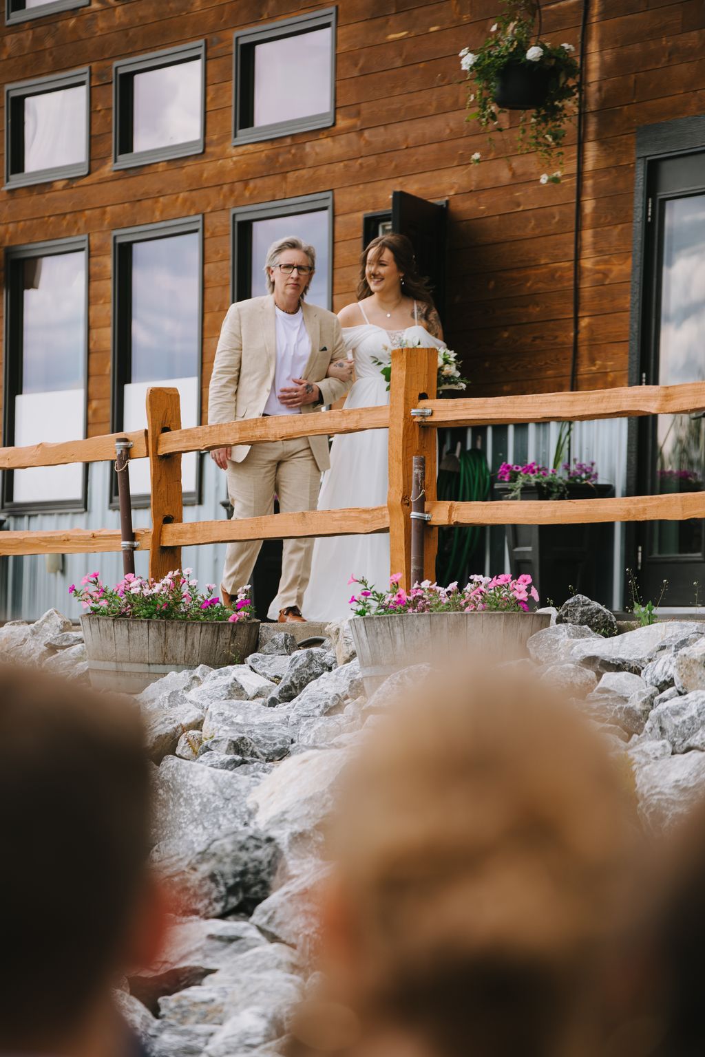 Groom in beige suit and bride in white dress lean against wooden railing with pink flowers and modern wooden home backdrop