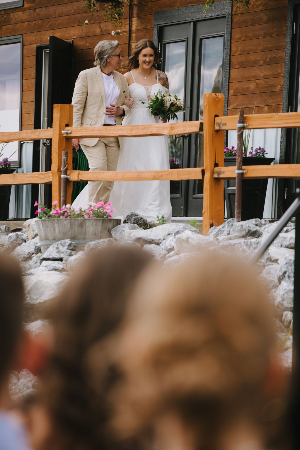 Bride in white dress holding bouquet walks arm-in-arm with woman in beige blazer past wooden fence in modern garden setting