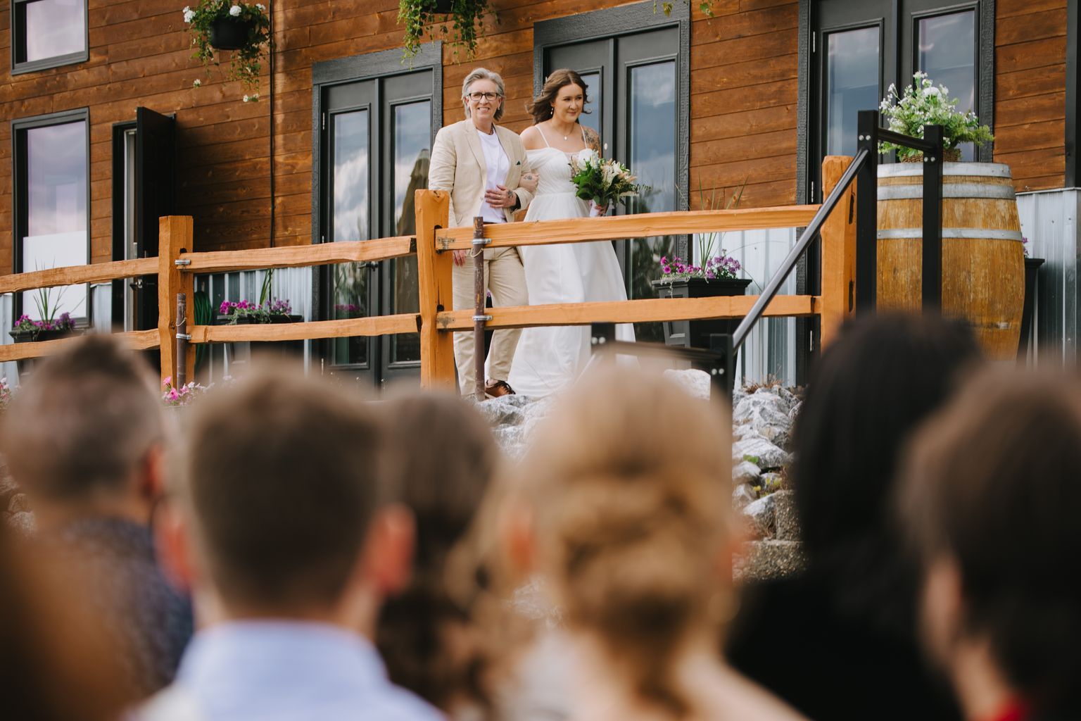 Bride in white dress with bouquet and older woman in beige cardigan stand at wooden railing outside modern home with dark windows