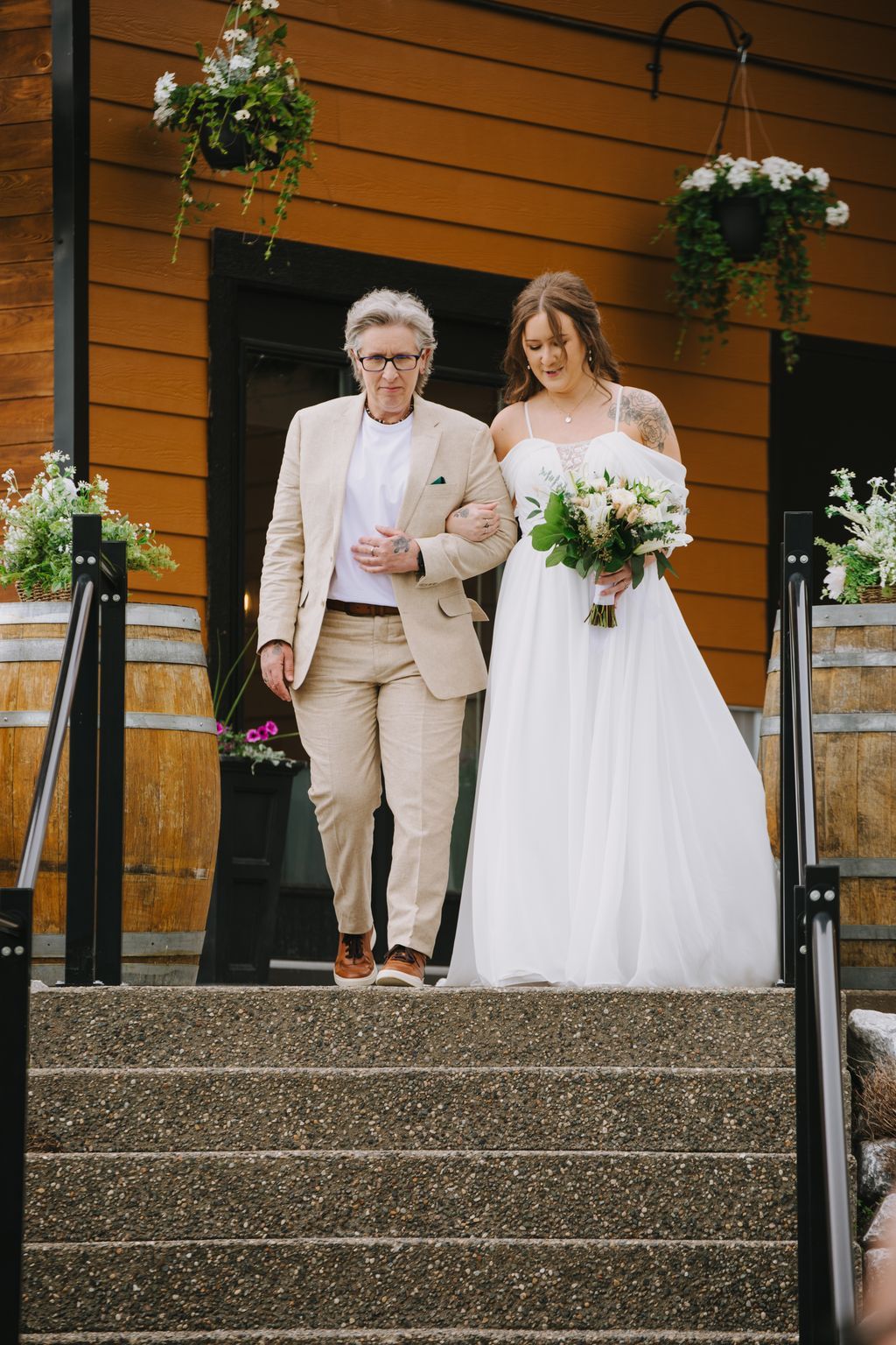 Bride in white dress with bouquet walks arm-in-arm with older man in beige suit down concrete steps outside modern wooden building
