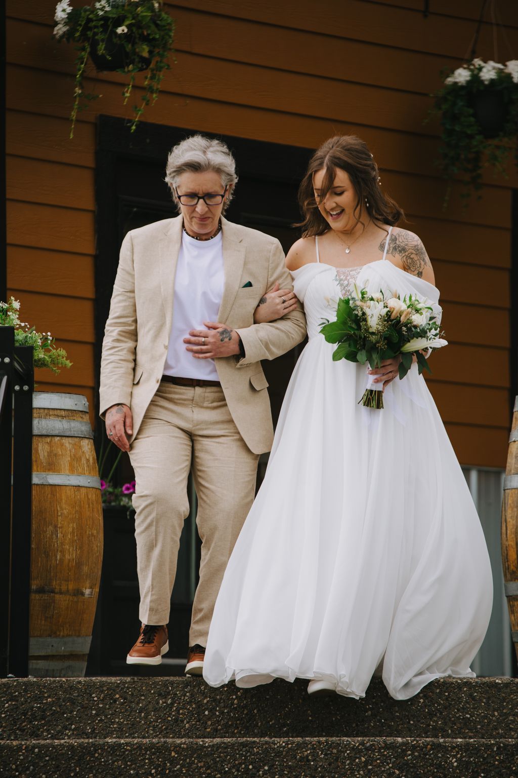 Bride in white off-shoulder gown and groom in beige suit walk arm-in-arm outside a wooden building, both smiling warmly