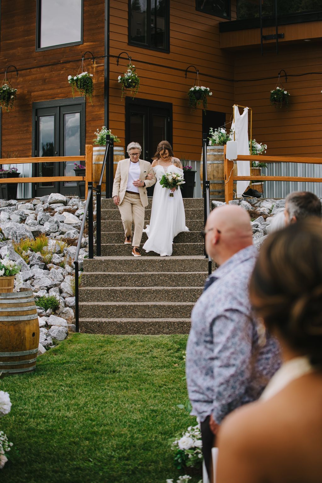 Bride and groom descend wooden steps at modern venue, bride in white dress holding bouquet, groom in tan suit, guests watching from lawn below