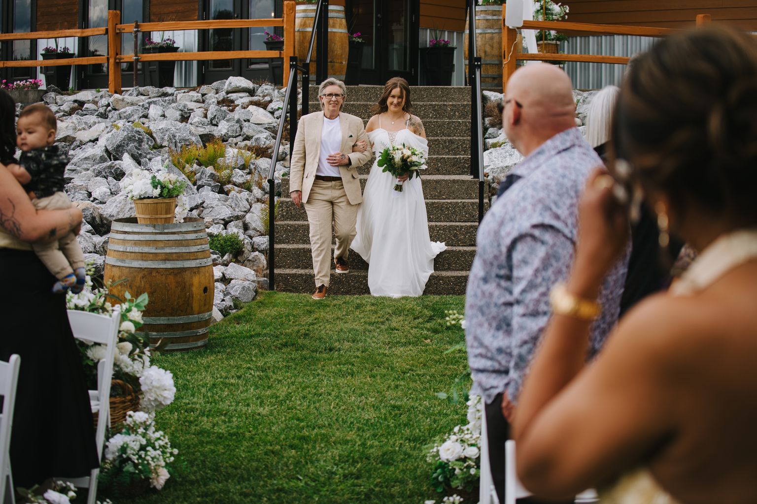 Bride and groom descend stone steps together, bride in white dress holding white bouquet, groom in cream suit, manicured lawn and modern building backdrop