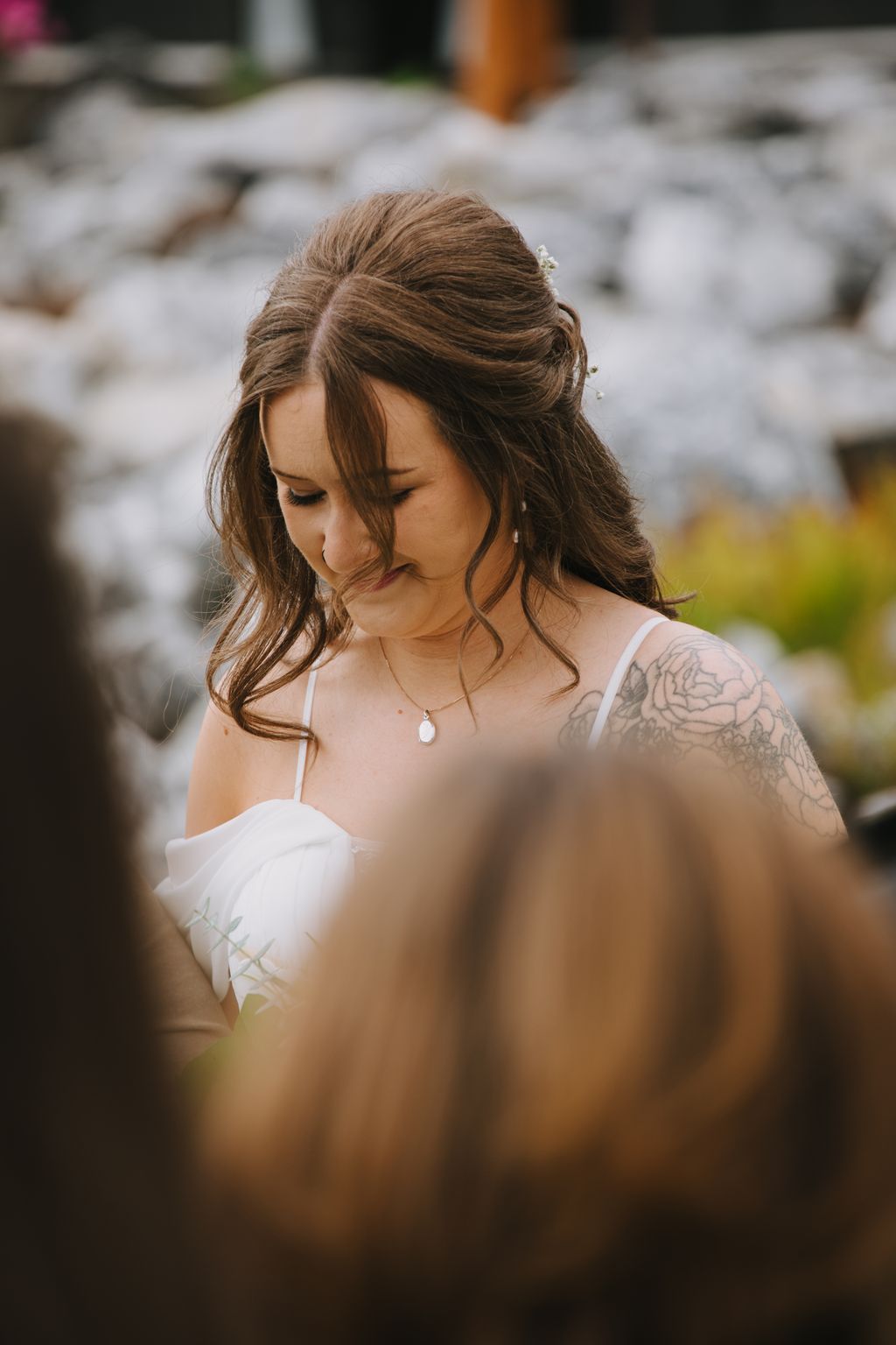 Bride with brown wavy hair and teardrop necklace gazes downward in soft natural light, wearing white dress against blurred outdoor background