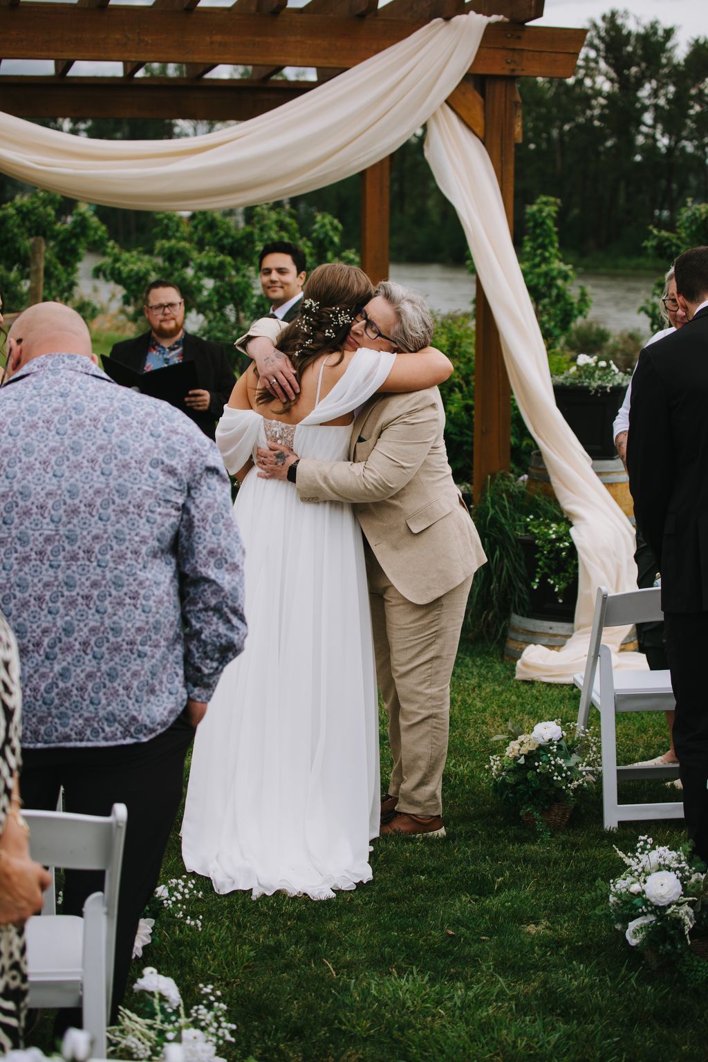 Bride in white dress embraces older woman under wooden pergola with flowing fabric draping at outdoor garden wedding ceremony