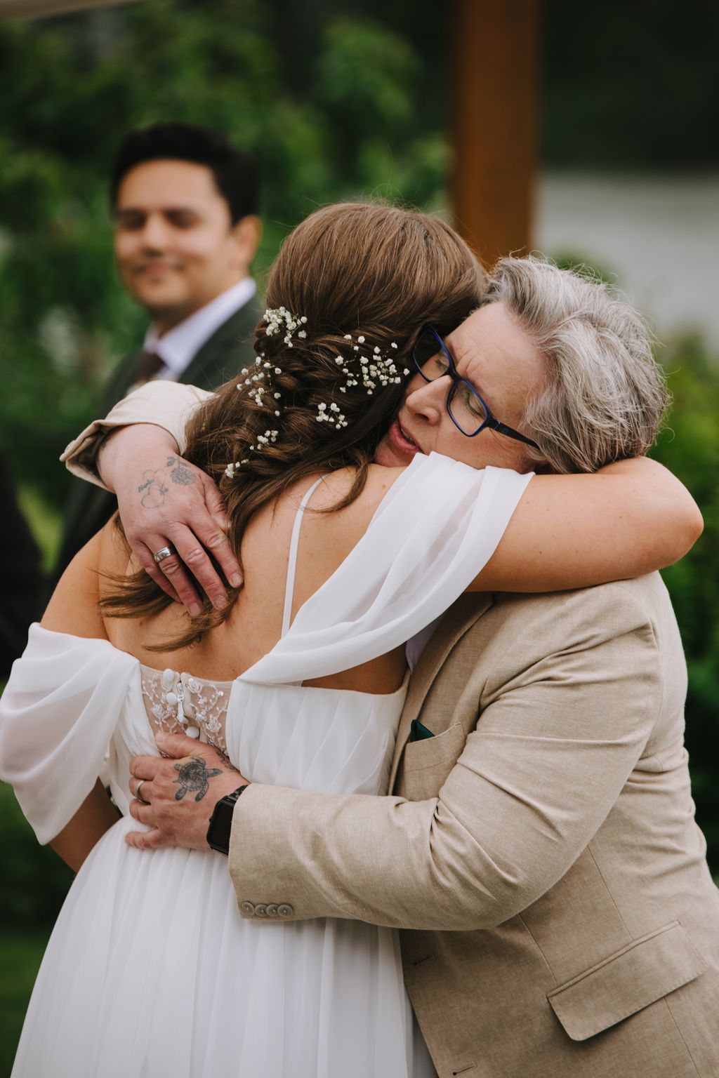 Groom in tan blazer embraces bride in white dress with baby''s breath in upswept hair, soft natural light