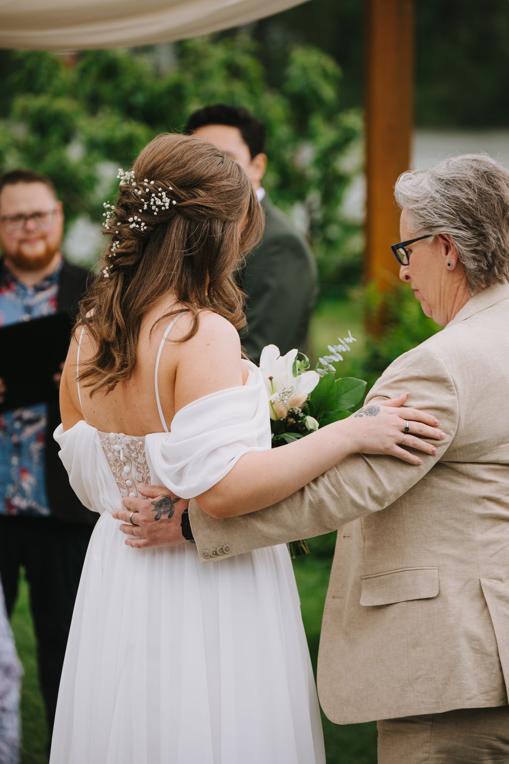 Bride in white off-shoulder dress with baby''s breath hair piece holds white bouquet while groom in beige suit embraces her from behind in garden setting