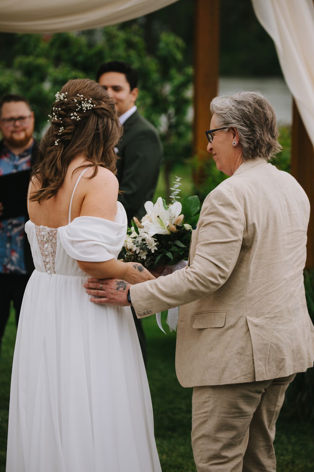 Bride in white off-shoulder dress with baby''s breath bouquet speaks with older woman in beige blazer at outdoor garden ceremony