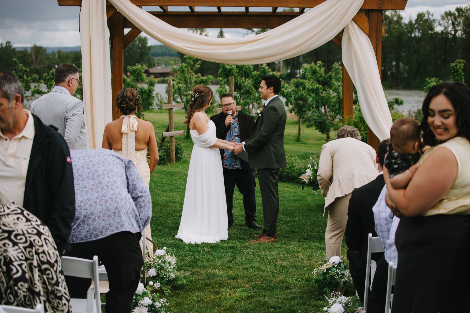 Bride in white dress and groom in olive suit exchange vows with officiant under draped pergola in vineyard ceremony