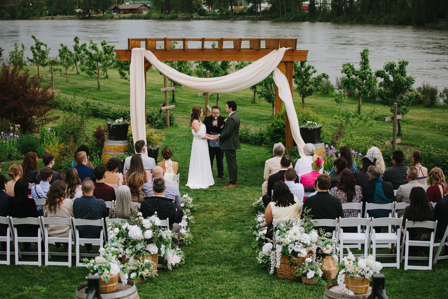 Bride and groom exchange vows under wooden arbor with draped fabric overlooking riverside lawn ceremony