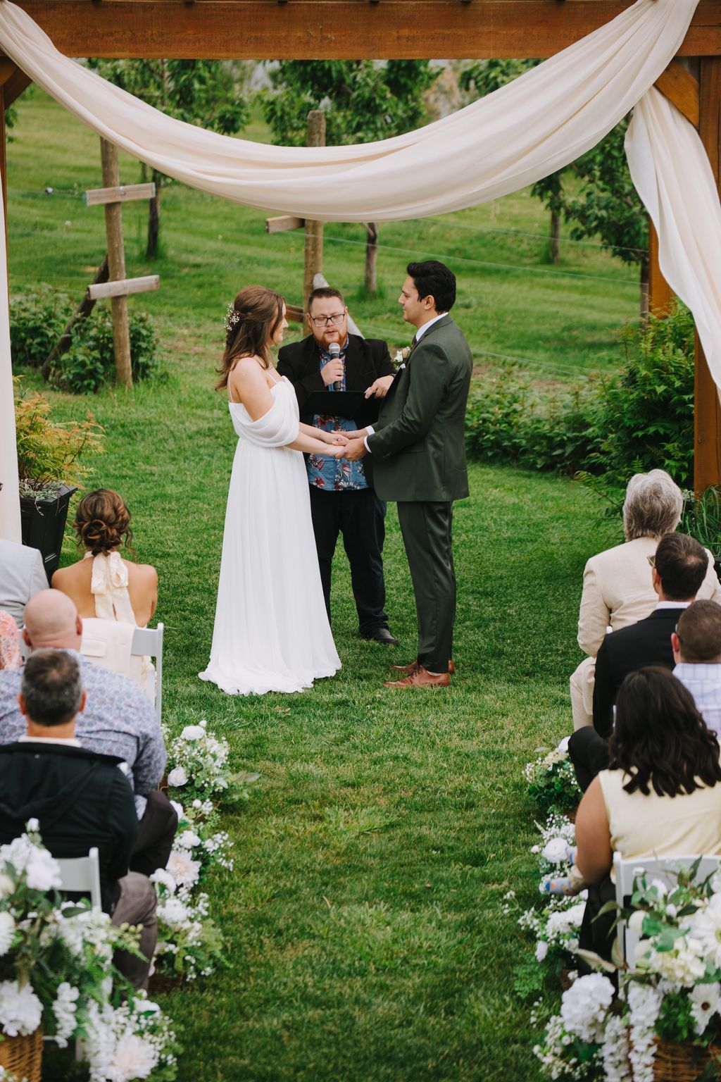 Bride and groom exchange vows under flowing white fabric canopy during outdoor garden ceremony with officiant