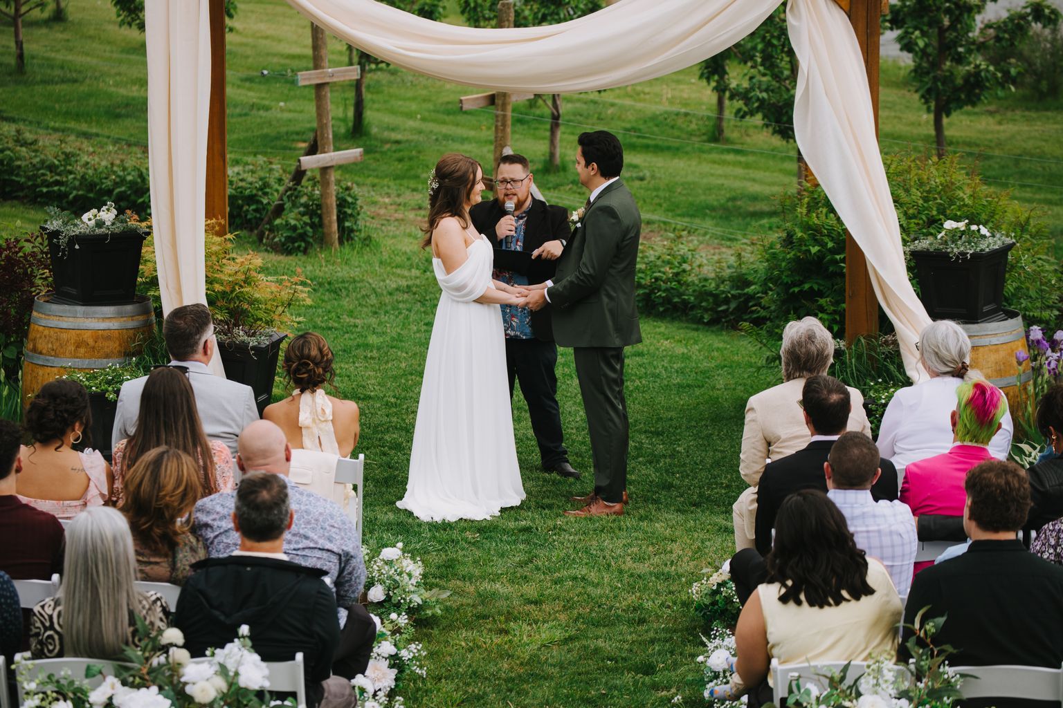 Bride and groom exchange vows under an elegant draped arbor while officiant holds microphone during outdoor garden ceremony