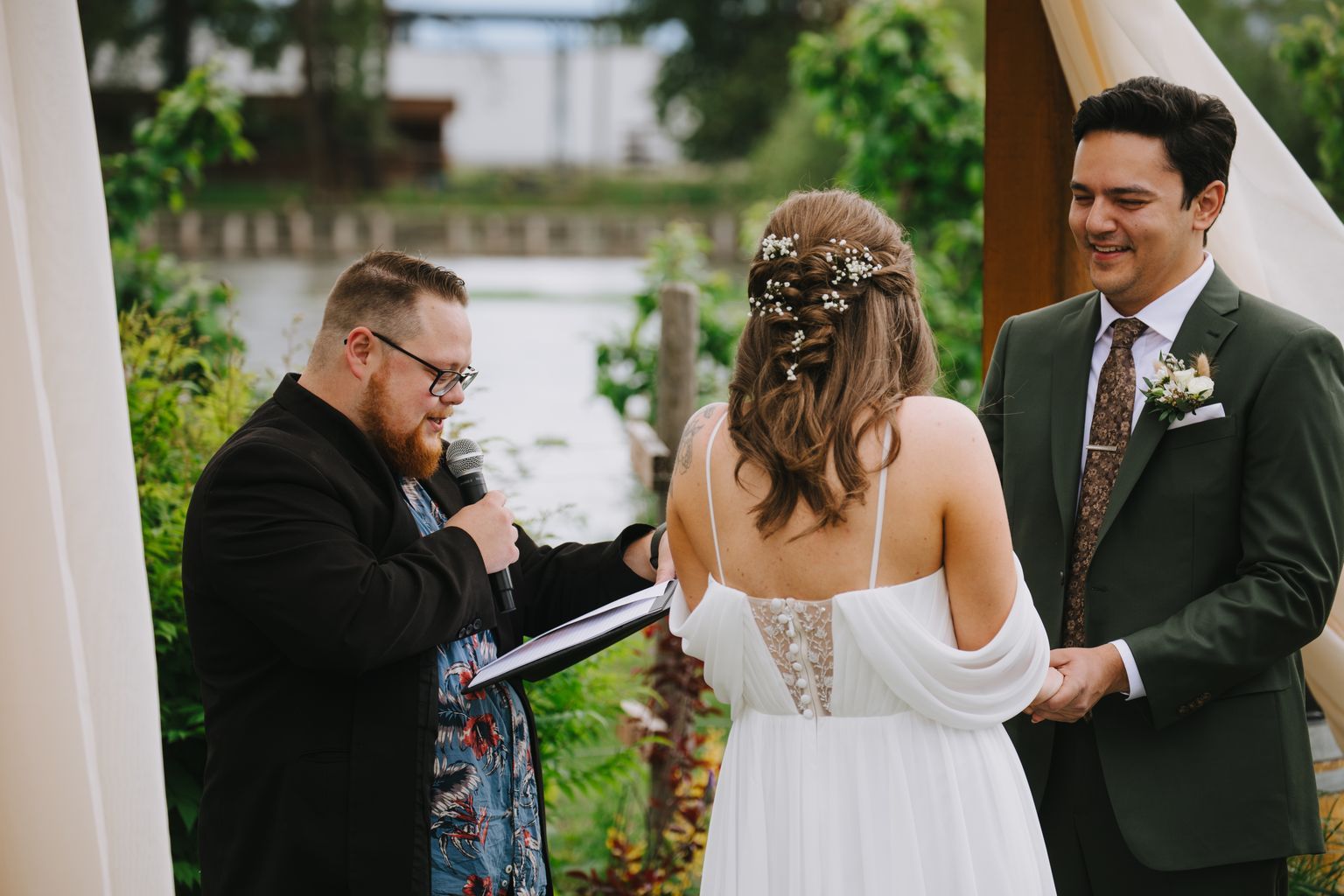 Bride in white lace dress with floral hair pins listens as officiant speaks during garden ceremony