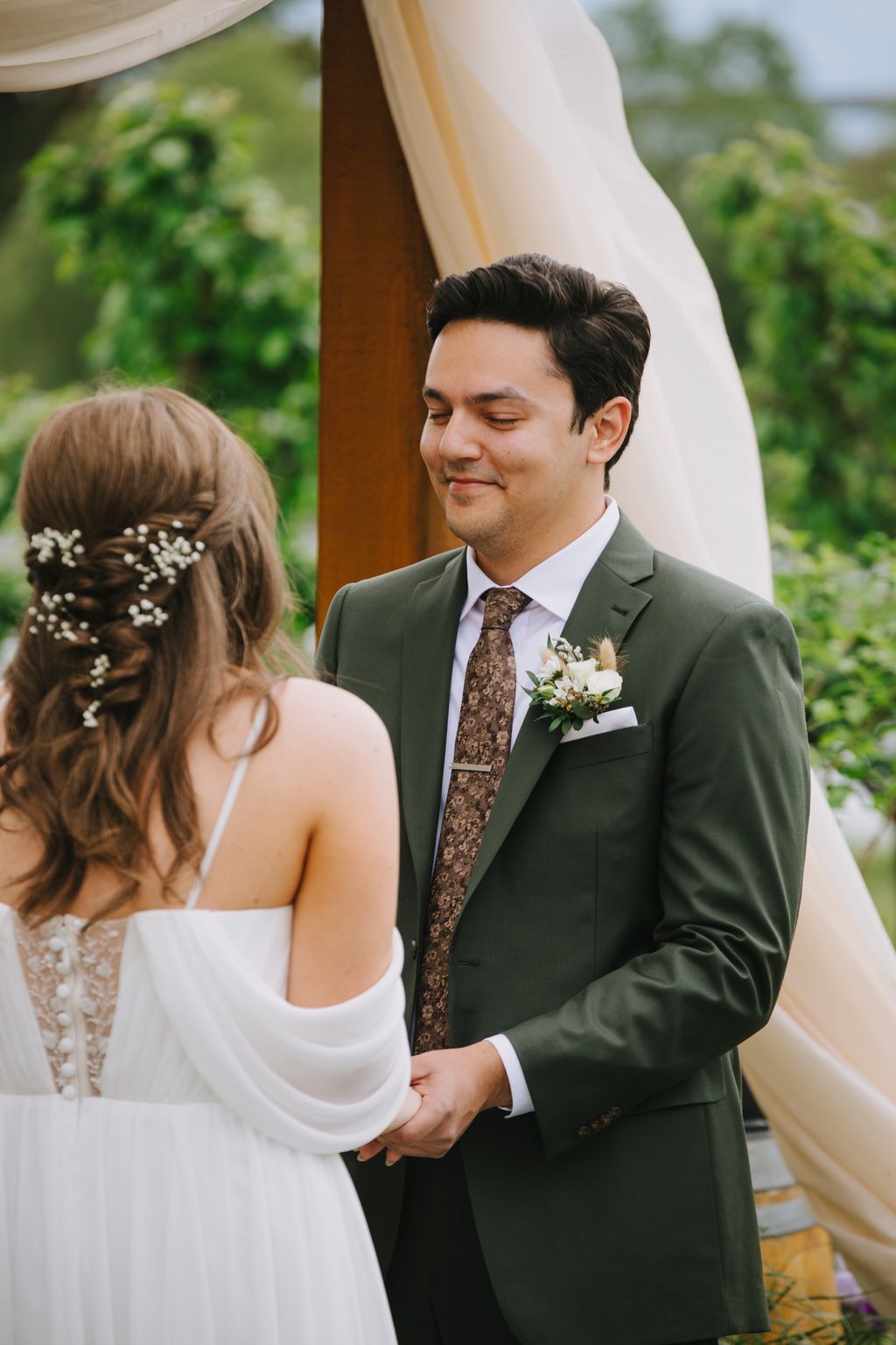 Groom in sage green suit and patterned tie holds bride''s hand under sheer white fabric drape, soft natural light and green foliage behind