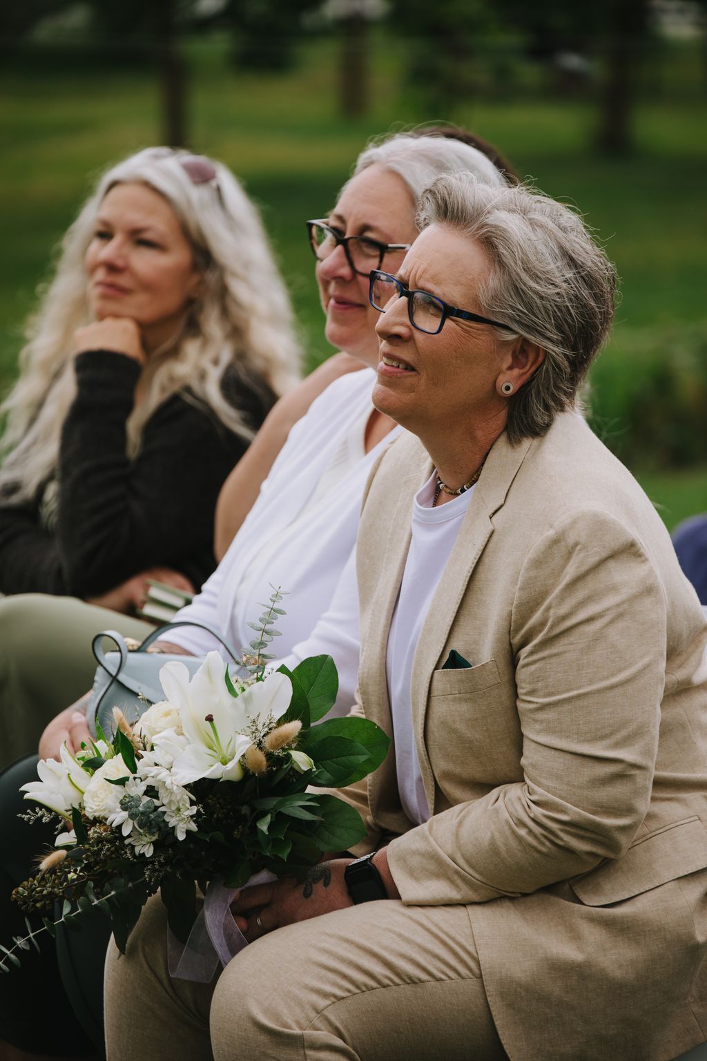 Two women in matching beige suits holding a white bouquet, smiling outdoors in natural light with green foliage behind them