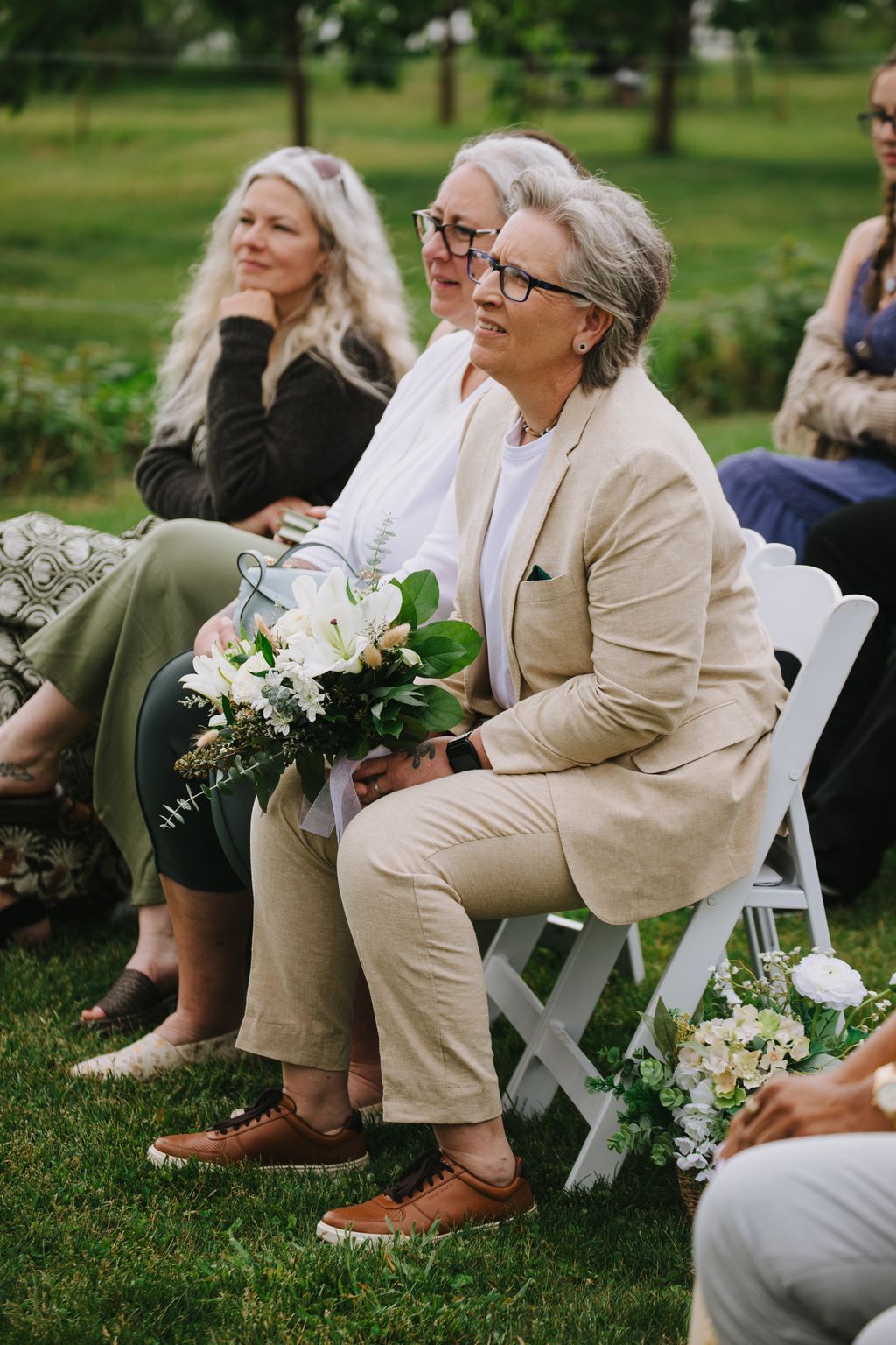 Three women in neutral tones seated outdoors holding a white flower bouquet, smiling in natural daylight at a garden ceremony