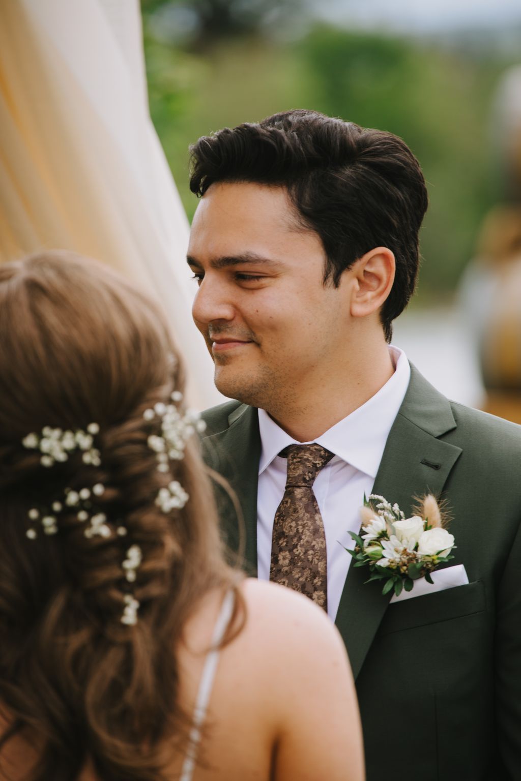 Groom in sage green suit and patterned tie gazes at bride during outdoor ceremony with soft natural light