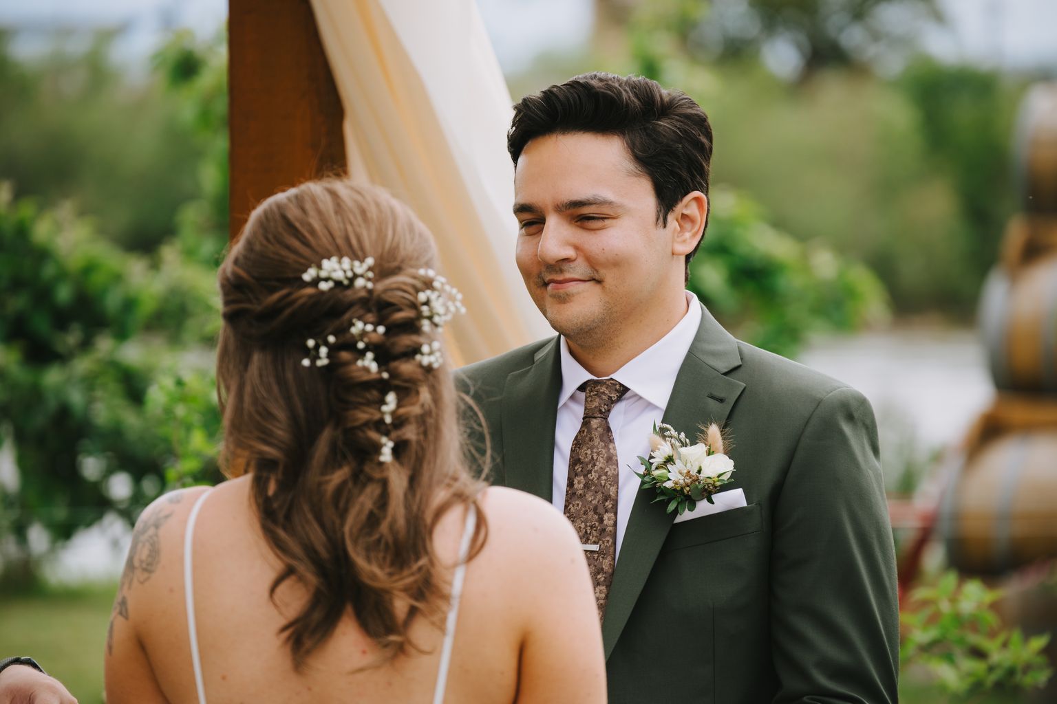 Groom in sage green suit with patterned tie gazes at bride in white dress during outdoor ceremony beneath sheer fabric