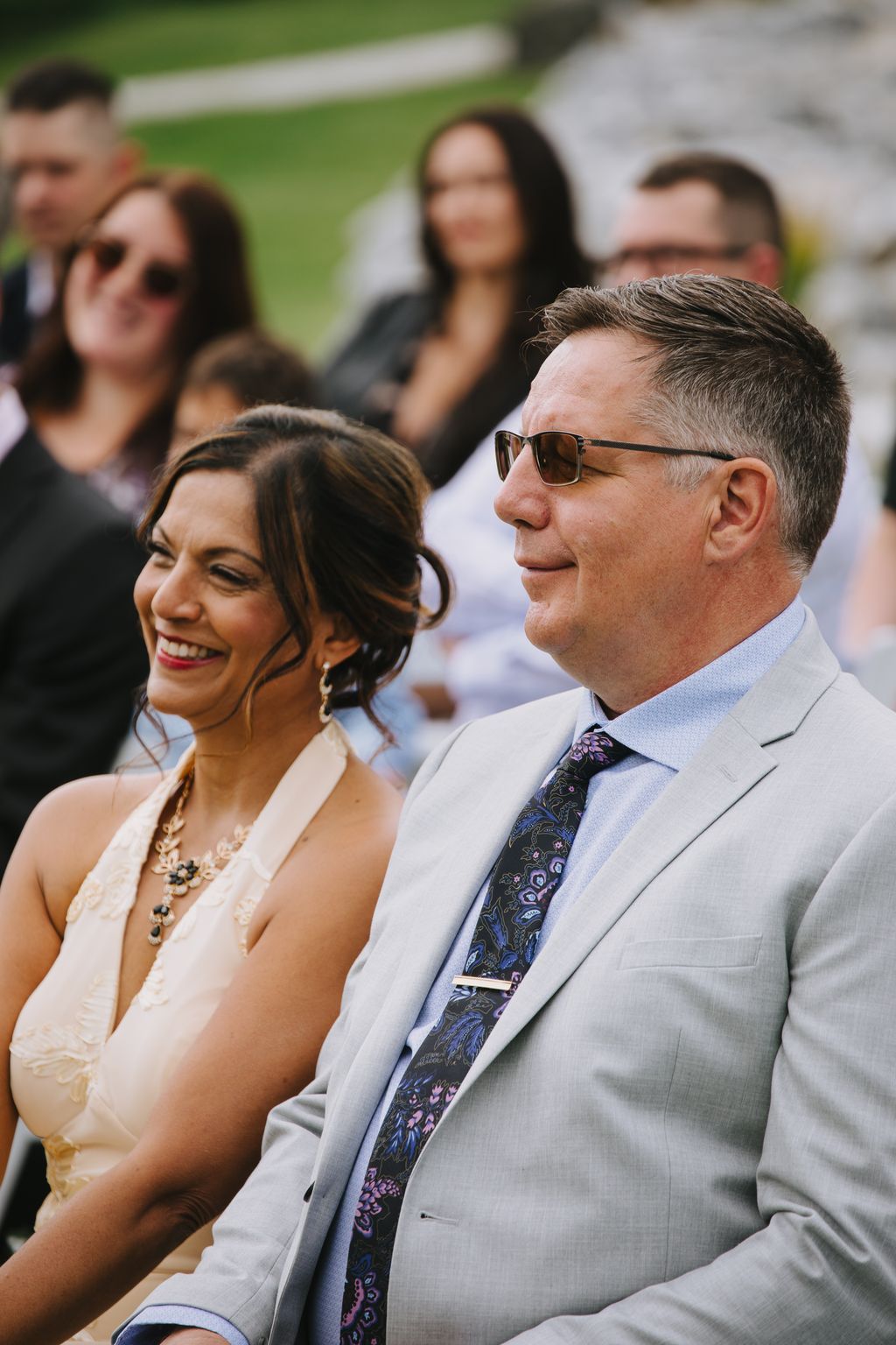 Groom in light gray suit and sunglasses smiles during outdoor ceremony with bride and guests blurred in background