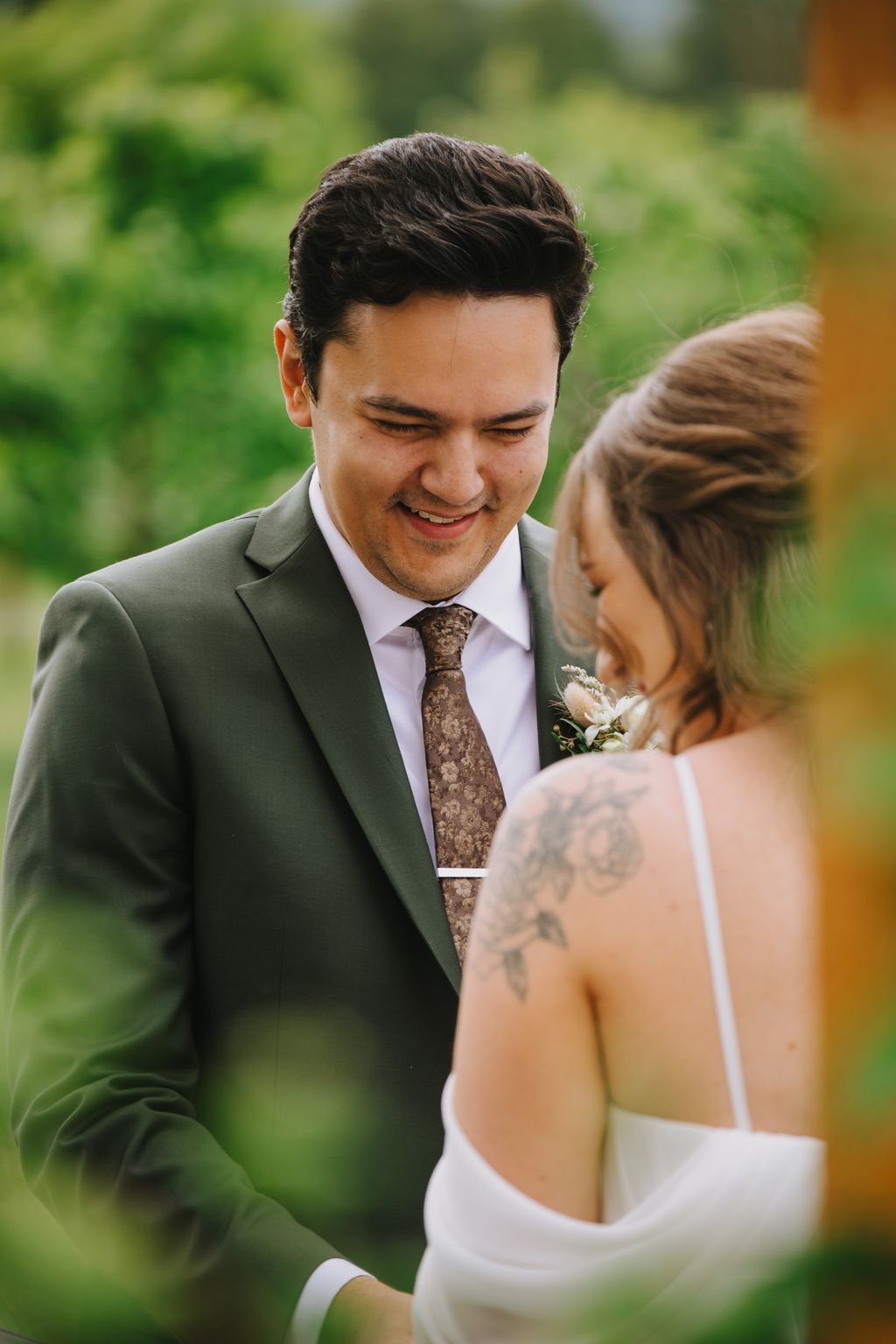 Groom in sage green suit and patterned tie smiles warmly at bride in white dress during outdoor ceremony with blurred green background