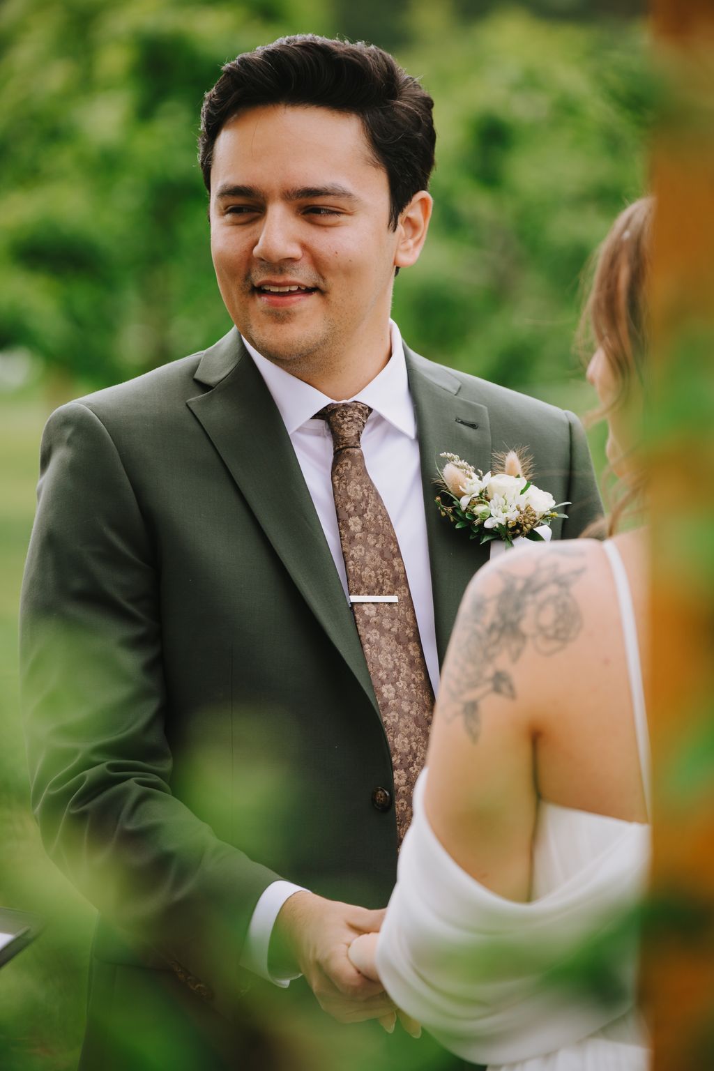 Groom in sage green suit and brown patterned tie smiles while holding bride''s hand outdoors with green background