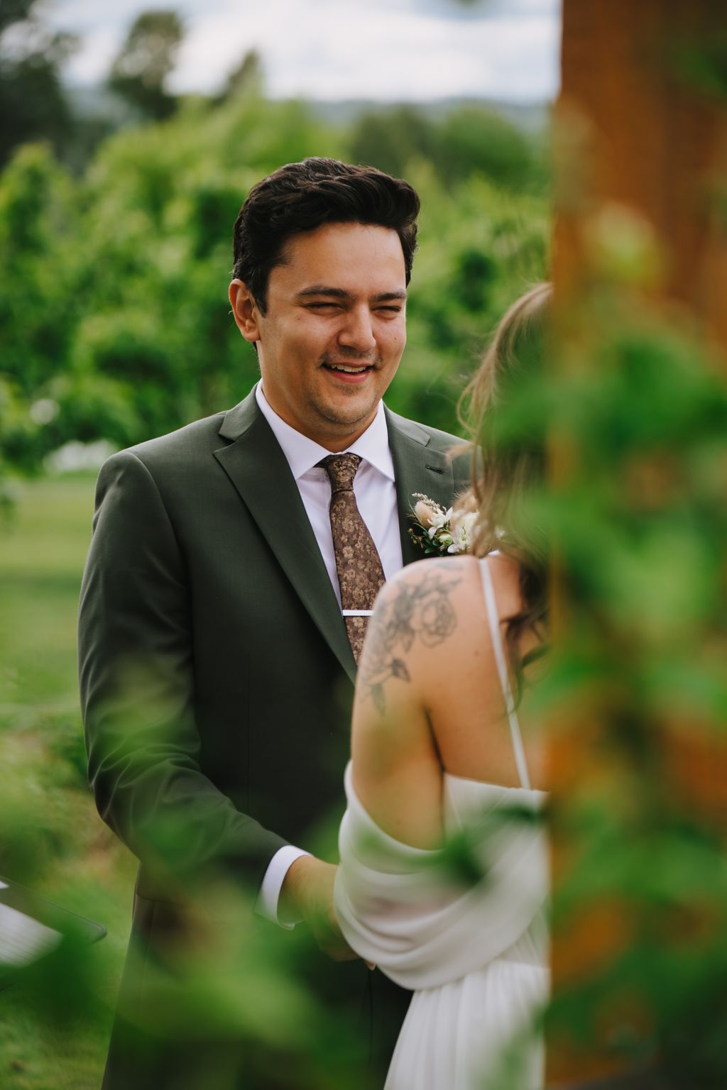 Groom in gray suit and patterned tie smiles at bride in white dress with visible tattoo, lush green landscape behind them