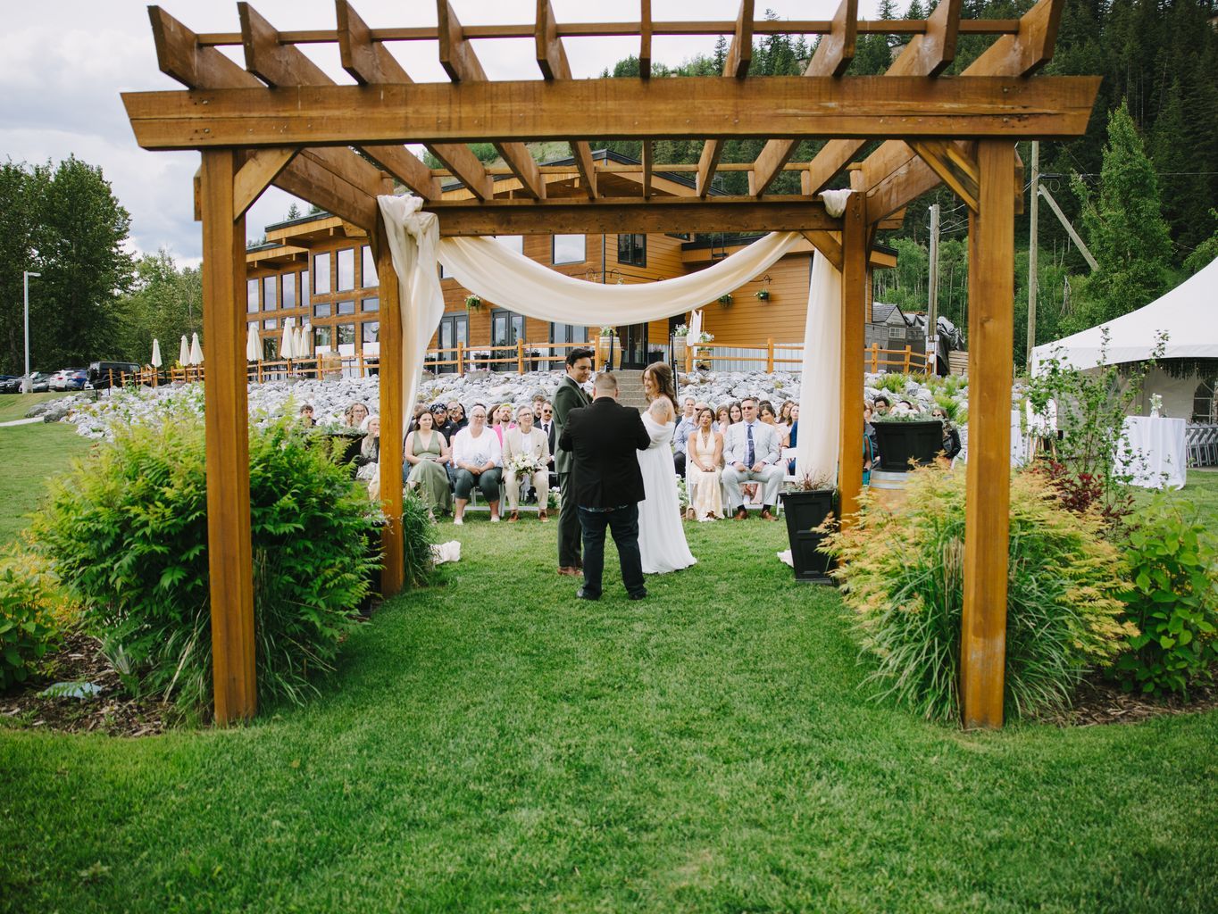 Bride and groom exchange vows beneath a wooden pergola with flowing fabric, surrounded by seated guests on a grassy lawn