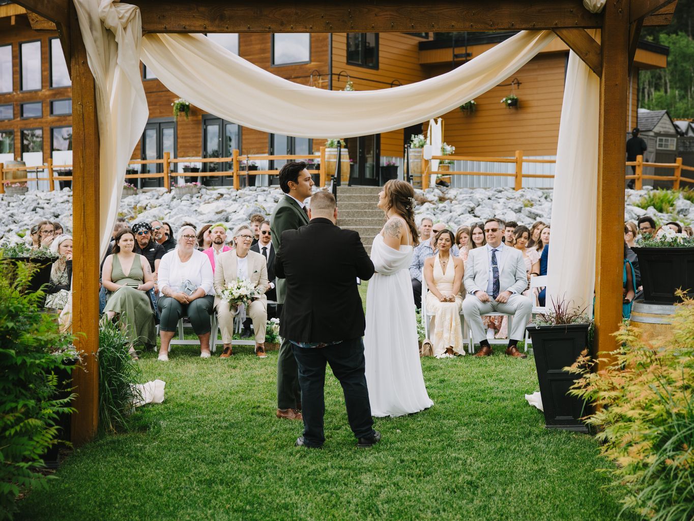 Bride in white dress and groom facing officiant during outdoor ceremony under draped fabric canopy with guests seated on grass