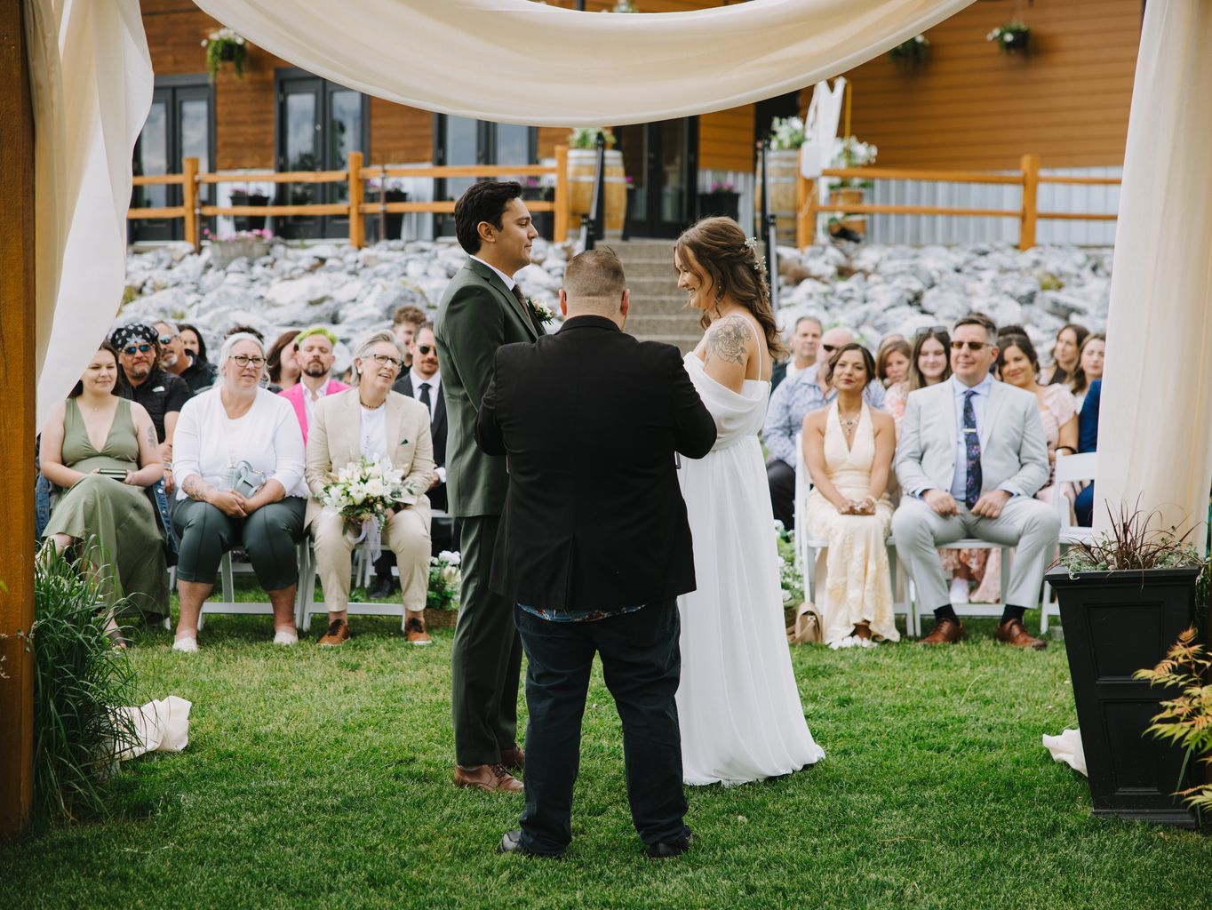 Bride and groom exchange vows with officiant during outdoor wedding ceremony under white draped arbor, guests seated on grass