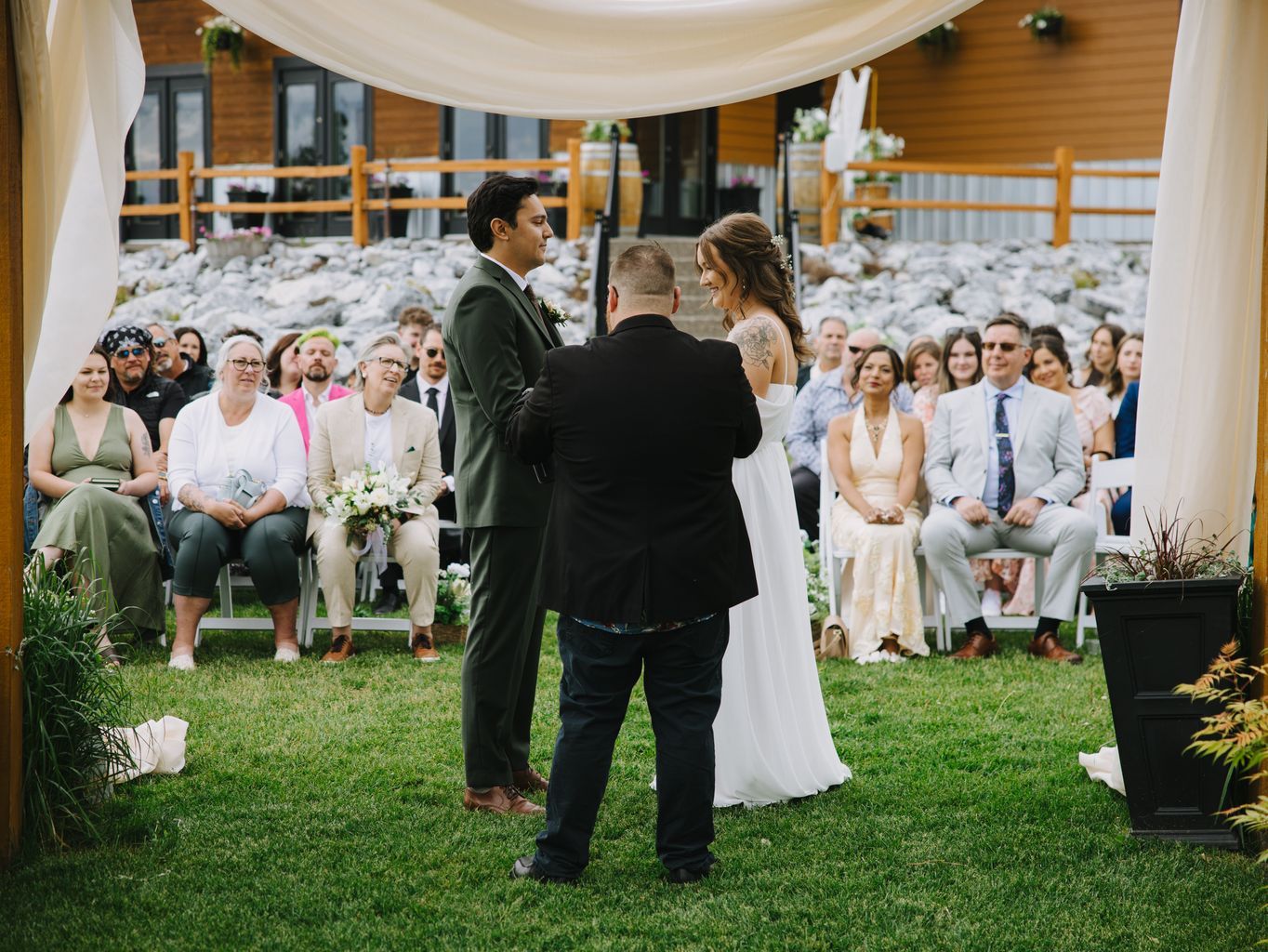 Bride in white dress and groom in sage green suit exchange vows with officiant under white tent at outdoor garden ceremony