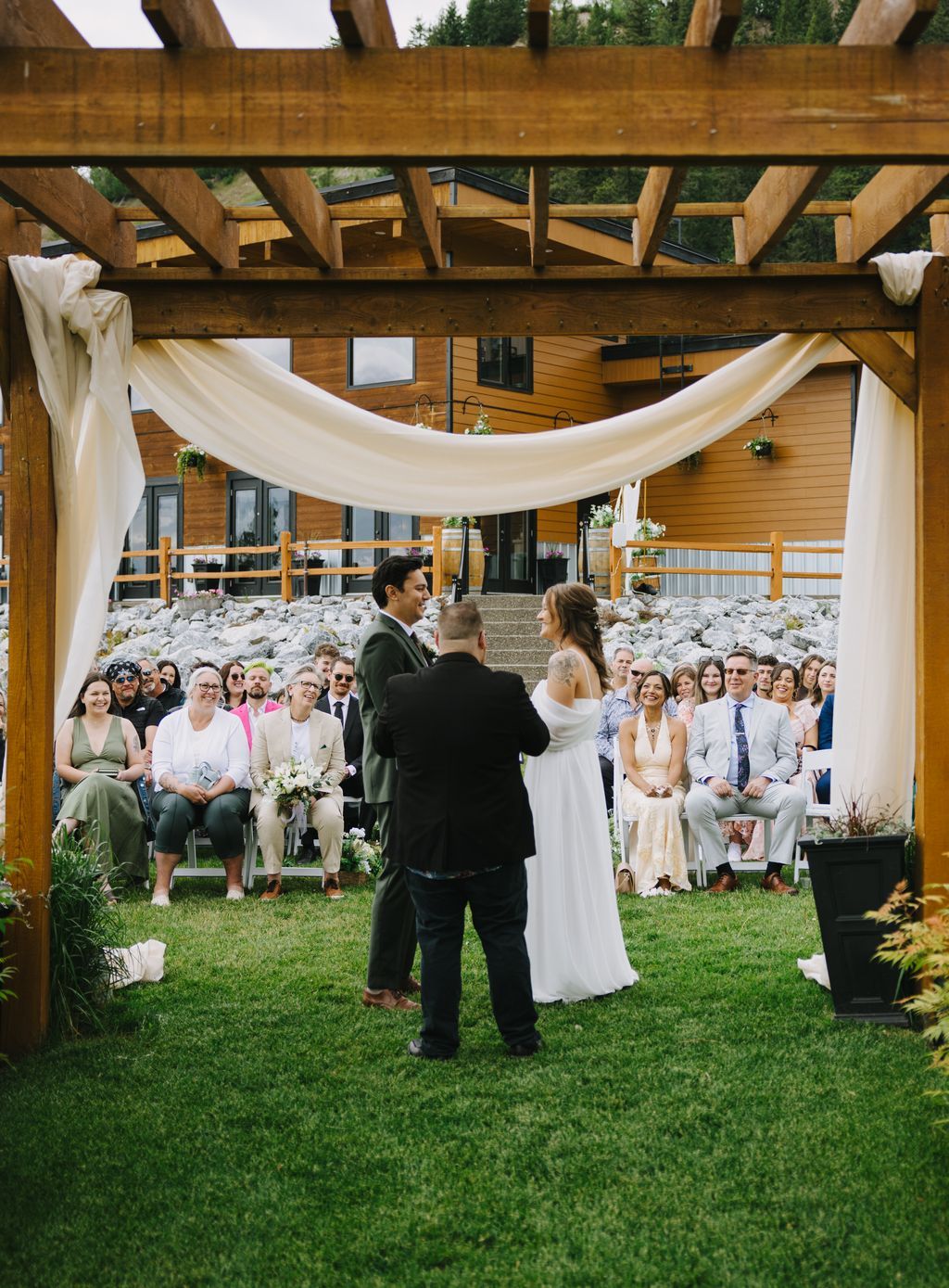 Bride and groom exchange vows during outdoor ceremony with guests seated on lawn beneath wooden pavilion