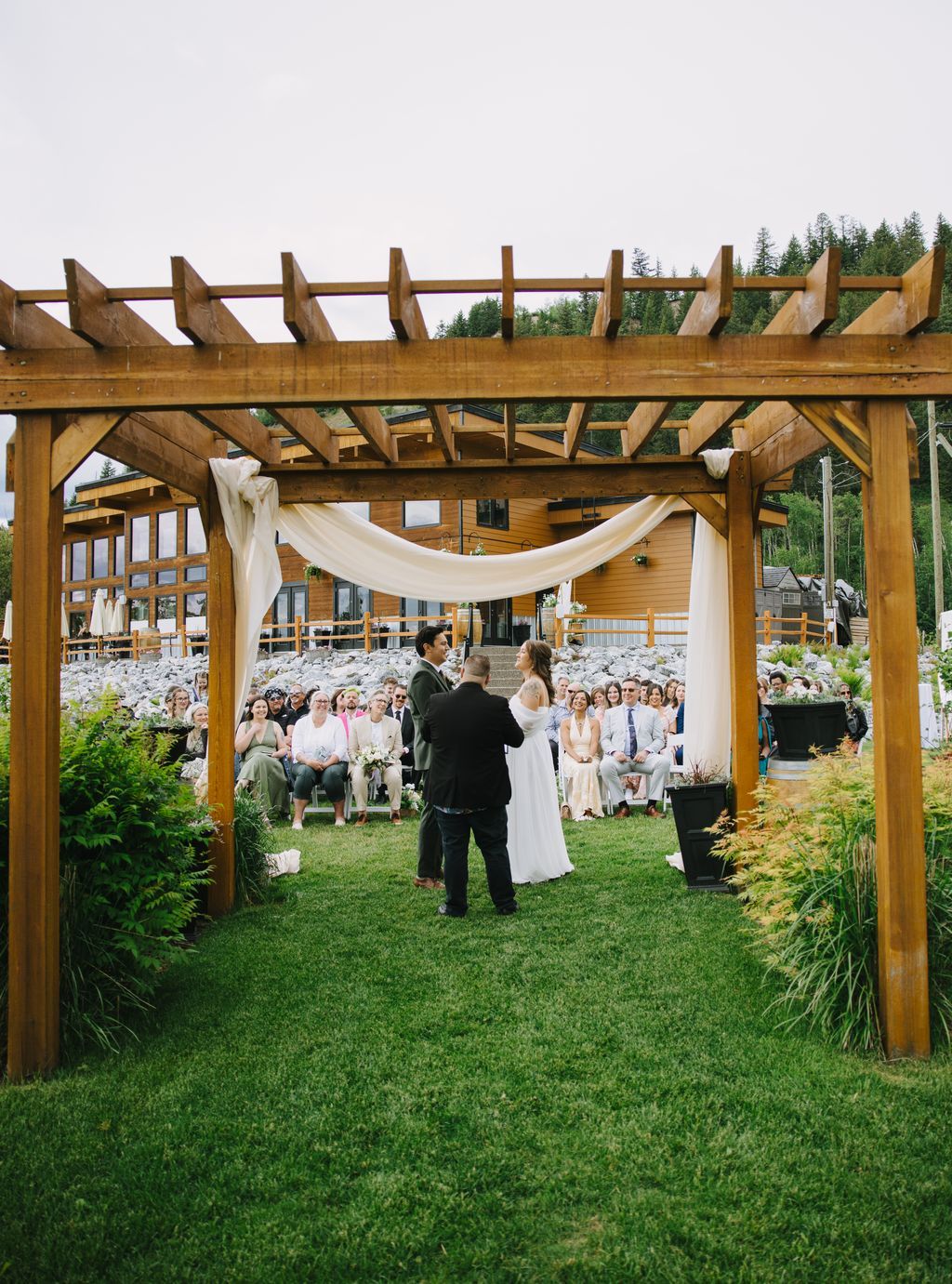 Bride and groom exchange vows under a wooden pergola with flowing fabric, surrounded by seated guests on a lush lawn