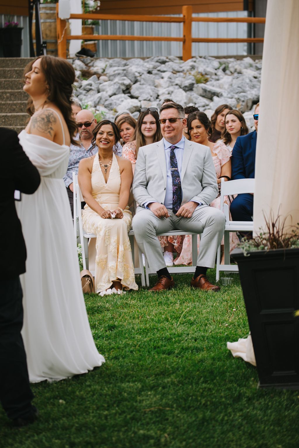 Couple seated together at outdoor wedding ceremony with guests in background, rustic stone wall and wooden fence behind them