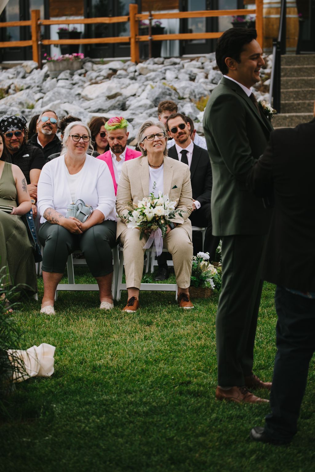 Bride in beige suit holding white flowers sits smiling during outdoor ceremony with guests and stone backdrop