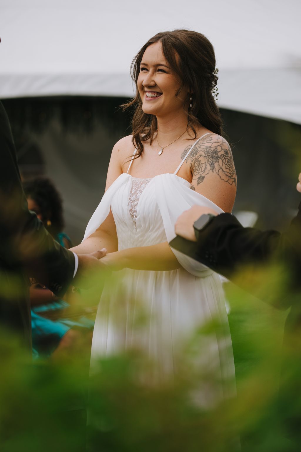 Smiling bride in white flowing dress with tattoo visible on shoulder, holding hands during outdoor ceremony with blurred green foreground