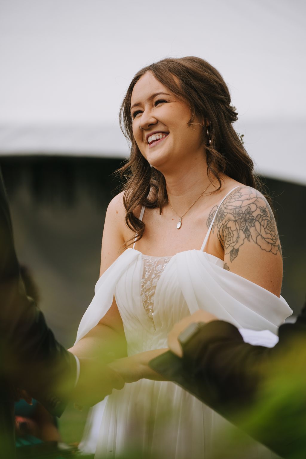Smiling bride in white off-shoulder dress with pendant necklace, outdoor natural light, soft green background