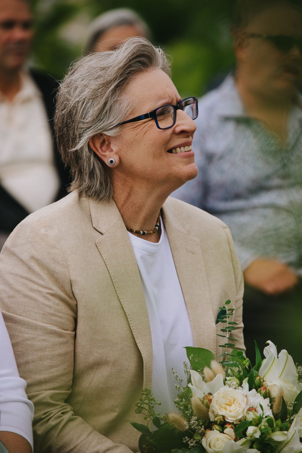 Woman in cream blazer and glasses smiles outdoors holding white flowers, soft green background