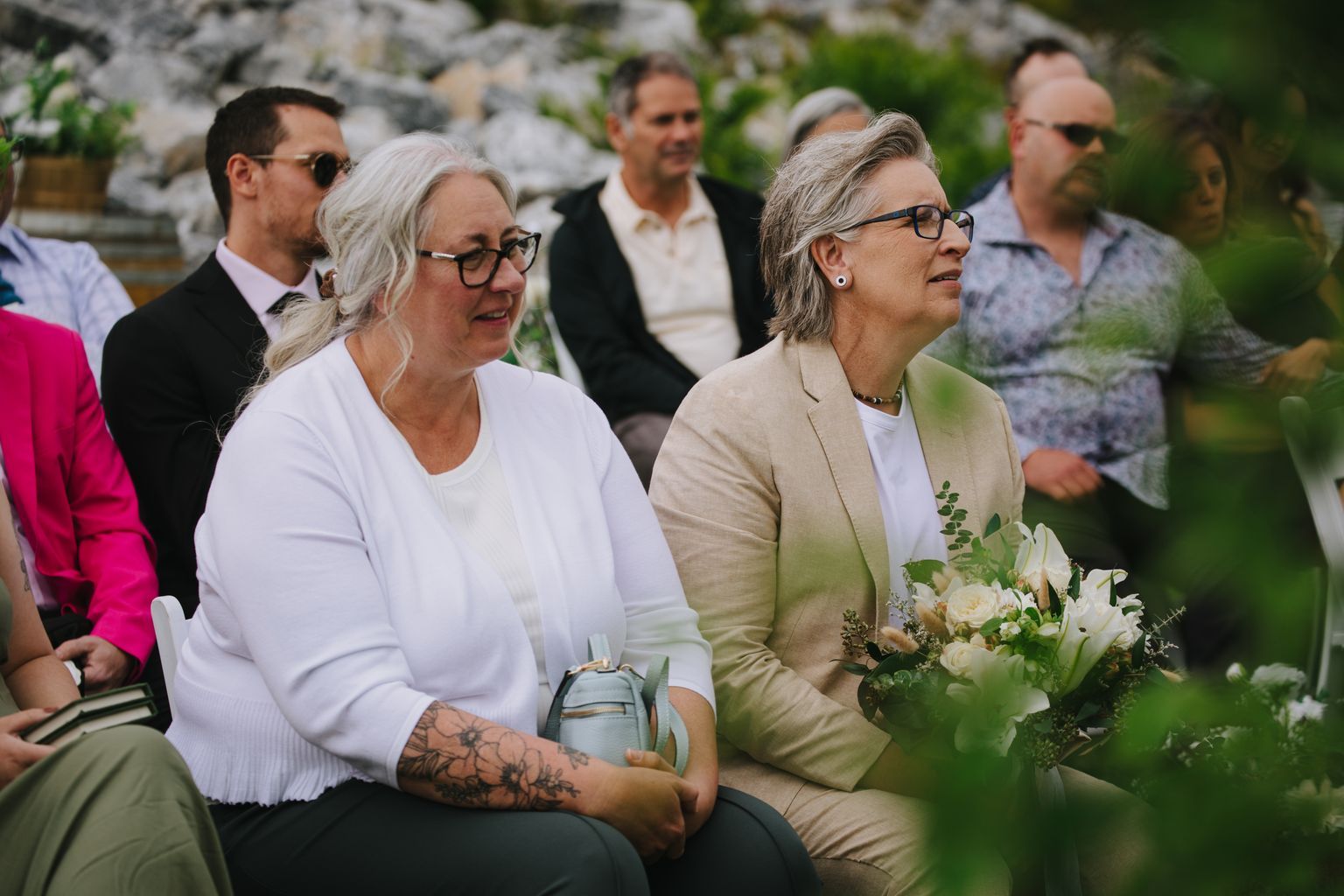 Two women sit together outdoors during a wedding ceremony, smiling warmly with stone and greenery in the background